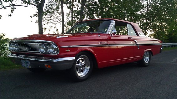 Red 1964 Ford Fairlane coupe parked on a road with trees and water in the background