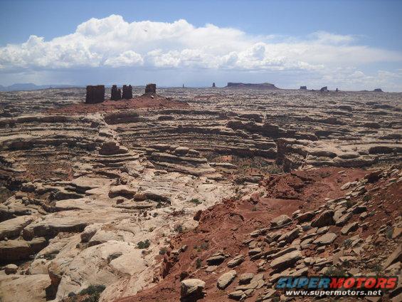 Desert landscape with layered rock formations under a cloudy blue sky. Supermotors.net logo in lower right