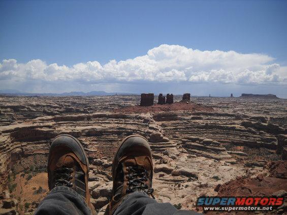 Person's legs and feet in brown hiking boots and dark pants sit on a rocky ledge overlooking a vast desert landscape with mesas and rock spires under a cloudy blue sky