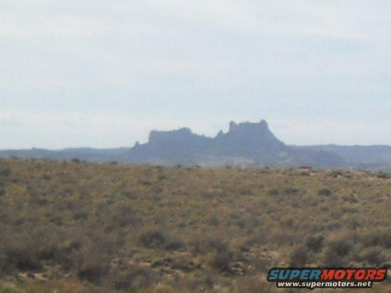 Desert landscape with large rock formations in the distance under a hazy sky. Dry brush covers the foreground