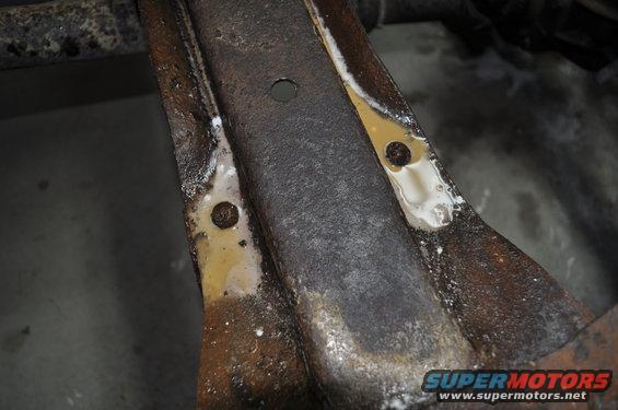 Close-up of rusted metal frame with yellow and white residue, showing bolt holes