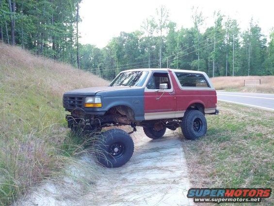 Two-tone lifted Ford Bronco with mismatched front end, parked on a dirt path next to a road