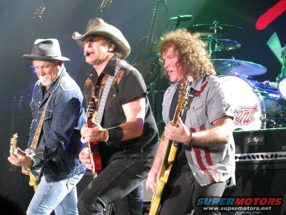 Three guys playing guitars on a stage with drums in the background