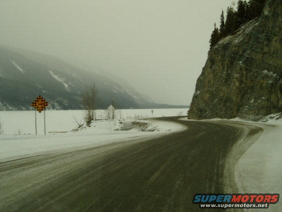 Winding snowy road next to a frozen lake with mountains in the background