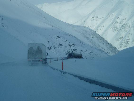 Two trucks driving on a snow-covered mountain road with steep, snow-covered slopes