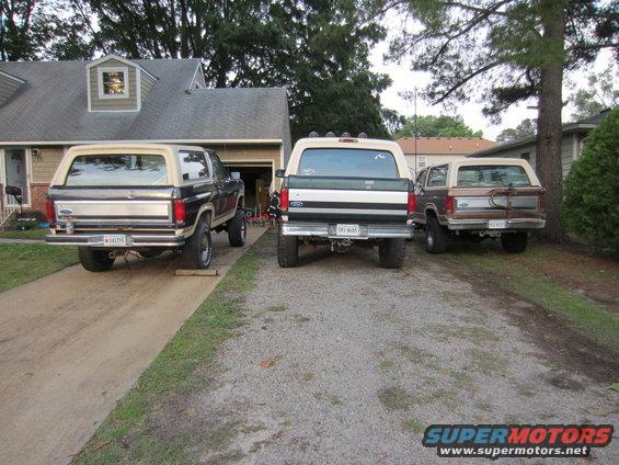 Three Ford Broncos parked in a driveway, two dark colored and one brown, all viewed from the rear
