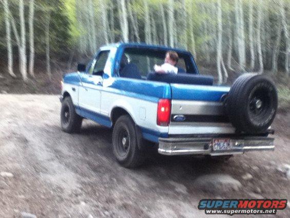 Blue and white Ford Bronco with spare tire on back driving on dirt road