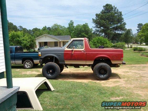 Lifted 1987 Ford Bronco, red and gray paint, large tires. Blue pickup truck parked behind it
