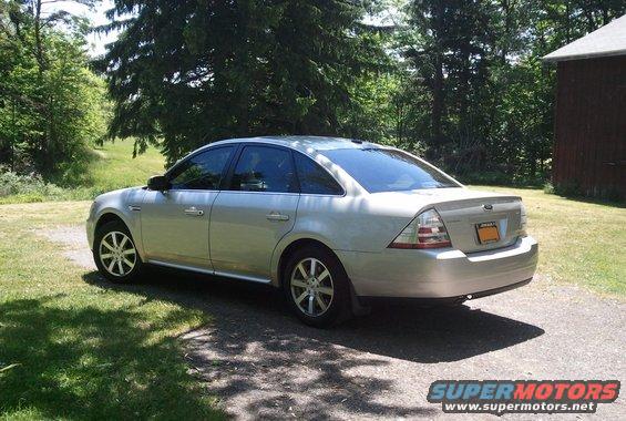 Silver sedan parked on a gravel driveway, rear 3/4 view. Trees and a barn are in the background