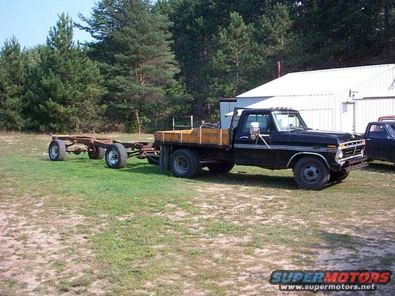 Black pickup truck with a wooden flatbed towing a trailer chassis. Another truck is partially visible to the right