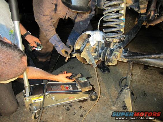 Mechanics working on the front suspension and brake assembly of a vehicle, which is supported by a floor jack and jack stand