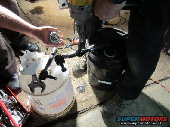 Mechanic drills into a pipe fitting assembly supported by a bucket, with tools nearby