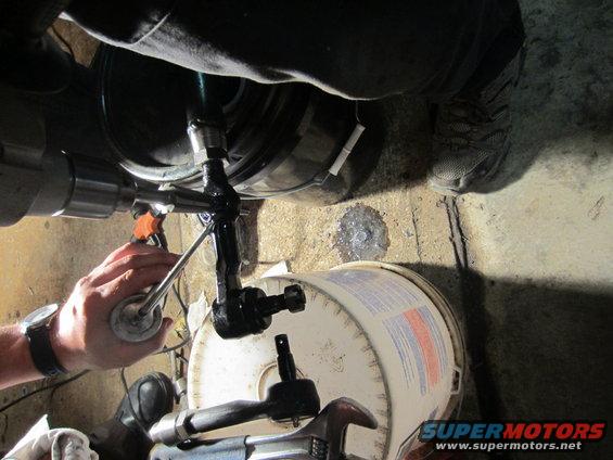 Steering linkage being worked on with a wrench and a spray can. A white bucket and concrete floor are visible