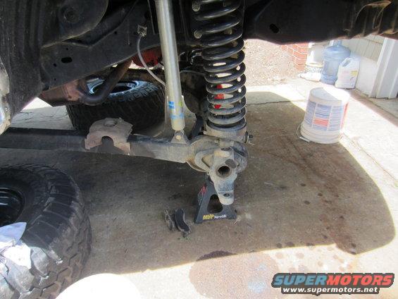 Close-up of a vehicle's front suspension assembly, including a shock absorber and coil spring, supported by a jack stand. A tire is visible to the left