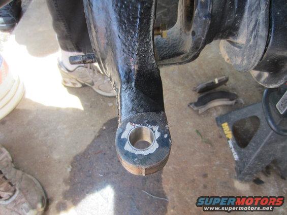 close-up of a vehicle's steering knuckle with a bolt and hole, brake pads and jack stand in background
