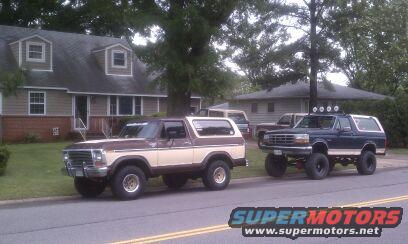 Brown and tan Ford Bronco from the early 1980s parked on the left, dark blue Ford Bronco from the 1990s lifted on the right