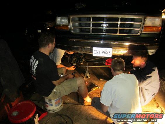 1992 Ford Bronco front end, dark green, with a chrome grille and orange turn signals. People are working on the steering components underneath
