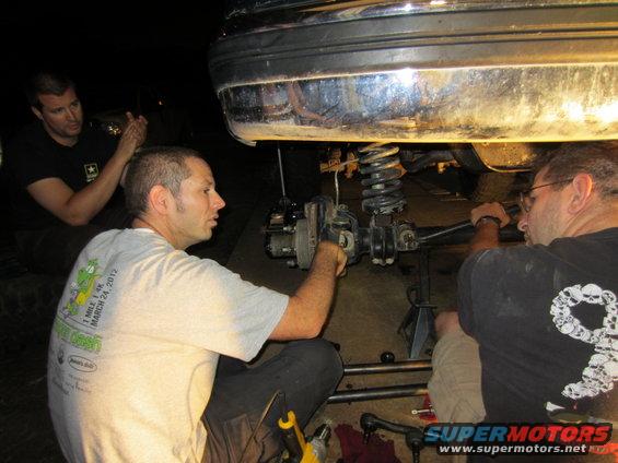 Undercarriage of a vehicle with suspension components visible, three men working on it