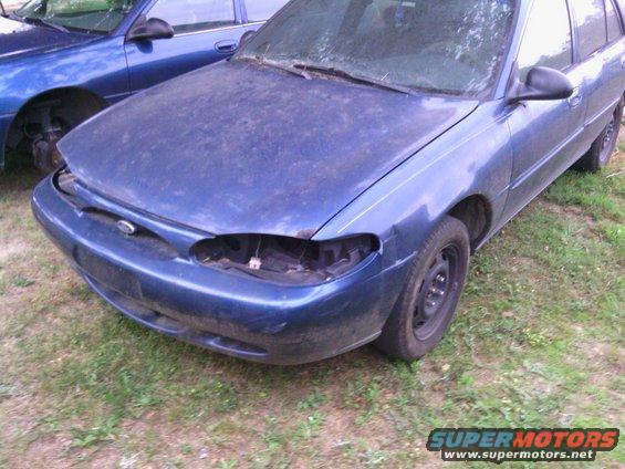 Blue sedan with missing headlights and dented fender, parked on grass