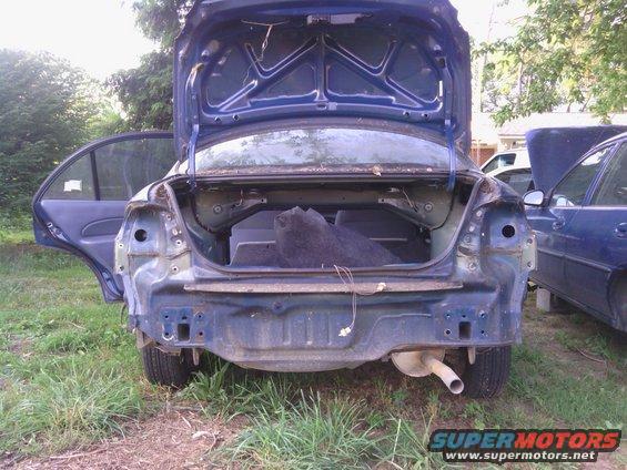Rear view of a damaged blue sedan, trunk open, bumper removed, exhaust visible. Another blue car is parked nearby