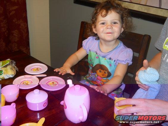 Child sits at a table with a pink toy tea set, including cups, plates, and a teapot