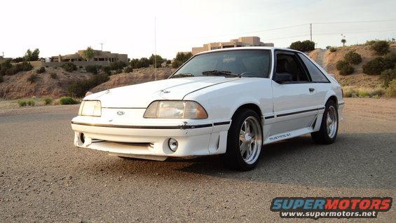 White Ford Mustang GT with aftermarket wheels, front bumper damage, and a visible "Mustang GT" badge on the side