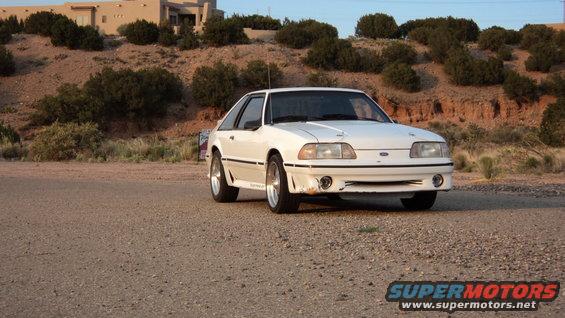 1989 Ford Mustang parked on a dirt road with a building and hills in the background