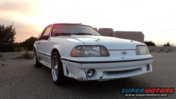 White Ford Mustang with aftermarket front bumper and hood pins, parked outside at sunset