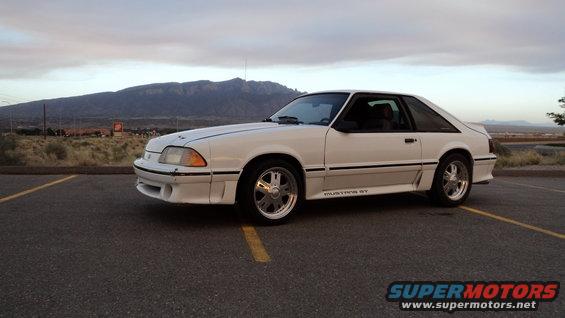 White Ford Mustang GT parked on asphalt with yellow lines, mountains in background