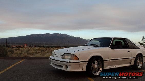 White 1989 Ford Mustang GT parked in a lot with mountains and cloudy sky