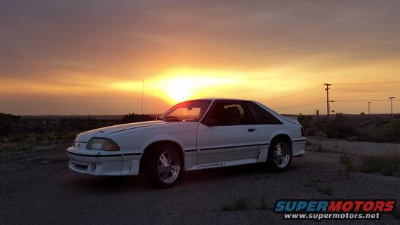 White 1987 Ford Mustang GT coupe with "MUSTANG GT" badging on the side, parked on a dirt road at sunset