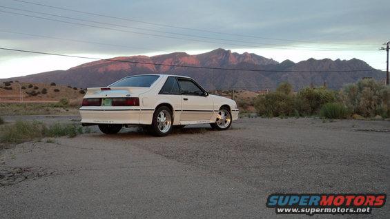 white fox body Mustang coupe parked on dirt lot, mountains in background