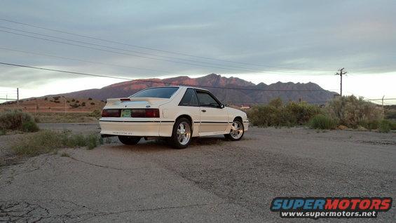White coupe with a spoiler parked on a gravel lot, mountains in the background