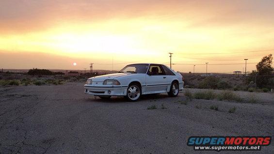 White 1987 Ford Mustang coupe parked on gravel with sunset in the background