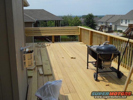 Wooden deck under construction with lumber, tools, and a black kettle grill. Houses and trees are visible in the background
