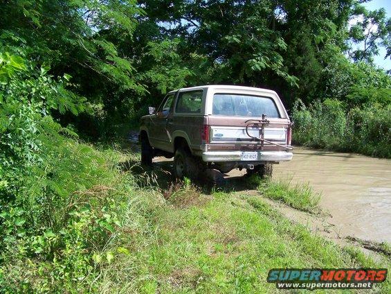 Brown 1985 Ford Bronco rear view, driving through muddy water with trees