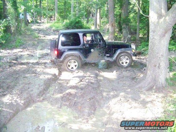 Black Jeep Wrangler driving through a muddy trail in the woods. Another vehicle is visible further up the path