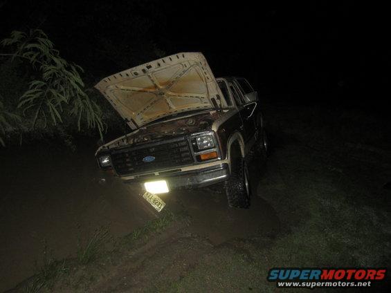 1980 Ford Bronco stuck in muddy ditch, hood open. License plate visible