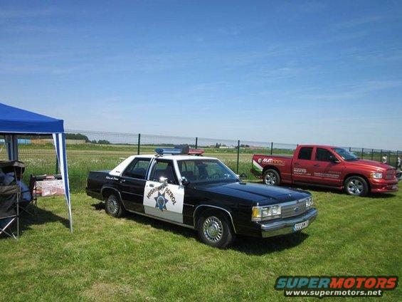 Black and white police car, 1991 Ford Crown Victoria, parked on grass with a red pickup truck behind it