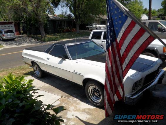 White Ford Ranchero with black top and hood, parked next to an American flag. Another vehicle is visible behind it