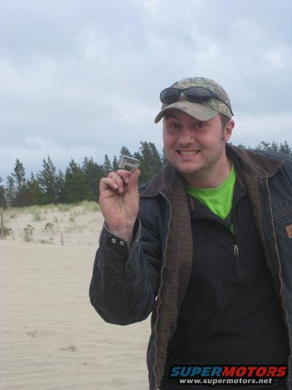 guy in a cap and jacket holds up a silver car emblem in front of sand dunes and trees