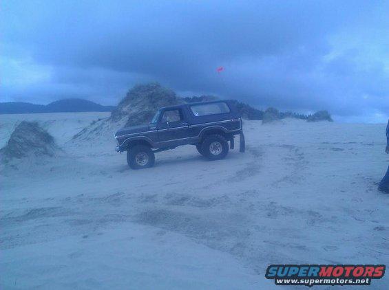 Dark blue Ford Bronco parked on a sandy beach with dunes and cloudy sky