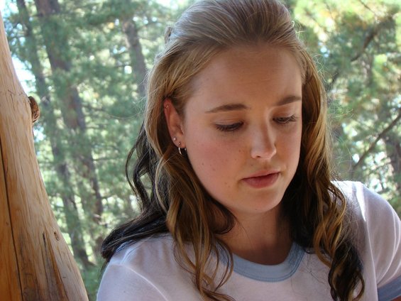 Young woman with long brown hair looking down, outdoors with trees in the background