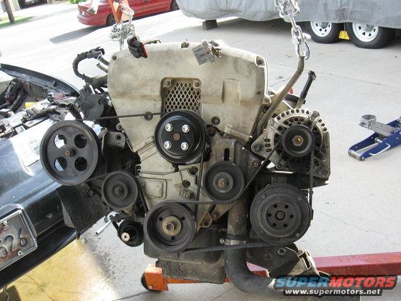 Engine assembly with multiple pulleys and belts, suspended by chains. A car fender and license plate are visible to the left