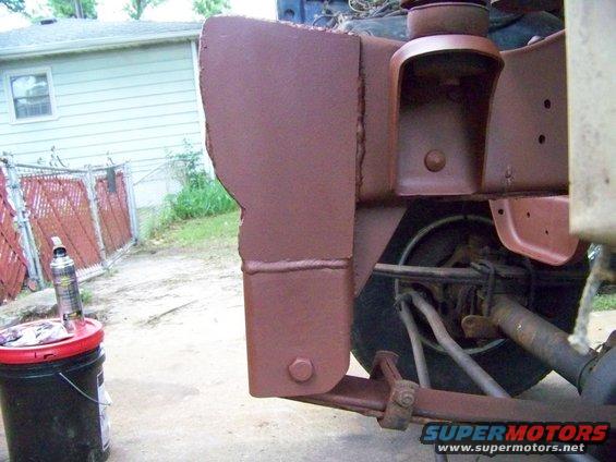 close-up of rusty vehicle frame and suspension parts, painted brown. A red bucket with a spray can sits nearby