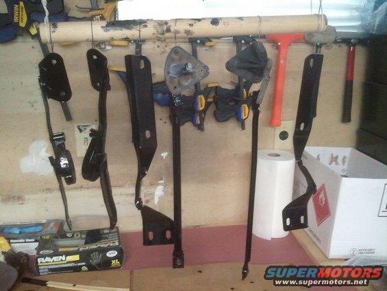 Several black metal car pedal assembly parts hanging on a wall, some with clamps attached. Box of gloves and paper towels nearby