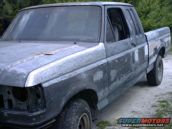 Gray Ford F-150 truck body in primer, driver's side front quarter view, showing body filler and missing headlight