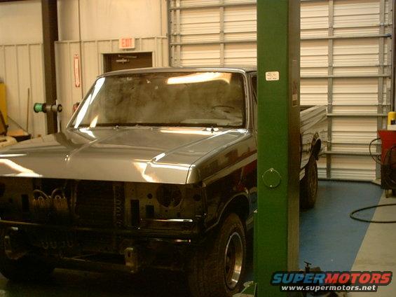Truck in garage, front end missing grille and bumper. Lifted on a green car lift