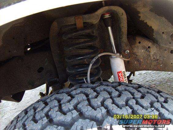 Close-up of a truck's front suspension with a coil spring and a white shock absorber labeled 'DOETSCH SHOCKS'. A large tire is in the foreground