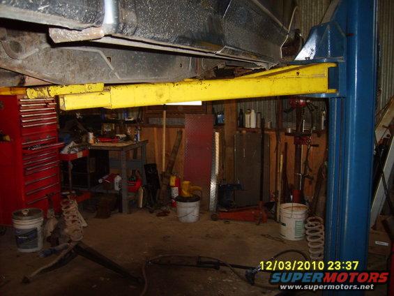 Underside of a vehicle on a lift in a garage, with tools and equipment visible
